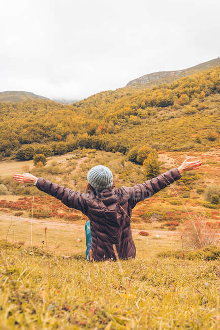 Vue arrière de dame en chapeau et veste de ski avec les mains levées assis sur le sol près des montagnes avec forêt à Isoba, Castille et Léon, Espagne — Photo de stock