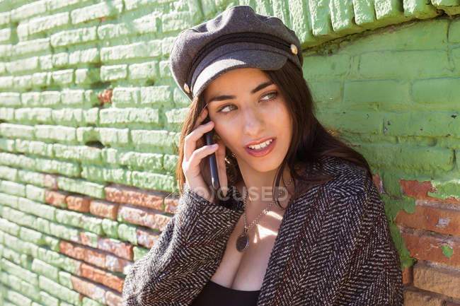 Sorrindo hispânico jovem mulher falando no telefone celular perto de parede de tijolo verde — Fotografia de Stock