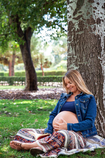 Young cheerful pregnant lady in casual wear holding belly and sitting on grass under wood in green park — Stock Photo