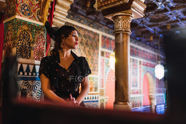 Side view of passionate young woman in dress sitting near table with chairs in luxury room of cafe decorated by mosaic — Stock Photo