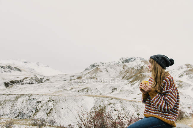 Vista lateral da jovem senhora em suéter e chapéu segurando caneca e olhando para colinas na neve em Pirinéus — Fotografia de Stock
