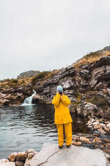 Mulher de chapéu e capa de chuva amarela em pé em pedras e disparando fenda de rio entre colinas de rocha em câmera em Isoba, Castela e Leão, Espanha — Fotografia de Stock
