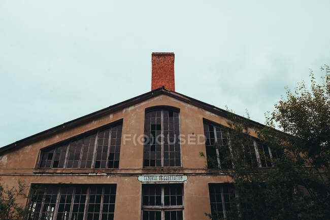 De baixo da fachada vermelha de construção quebrada envelhecida com janelas grandes e chaminé perto de árvores e céu nublado — Fotografia de Stock