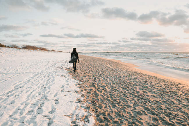На задньому плані жінка в пальто і капелюсі з рюкзаком йде на піщаний берег біля снігу і води в Клайпеді, Літуанія — стокове фото