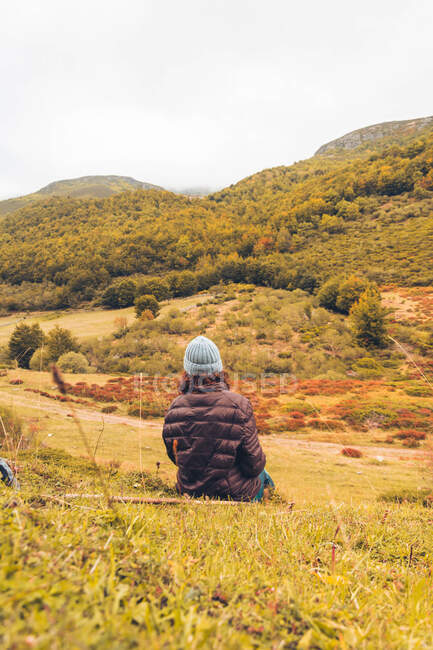 Vista trasera de la señora de sombrero y chaqueta de esquí sentada en el suelo cerca de las montañas con bosque en Isoba, Castilla y León, España - foto de stock