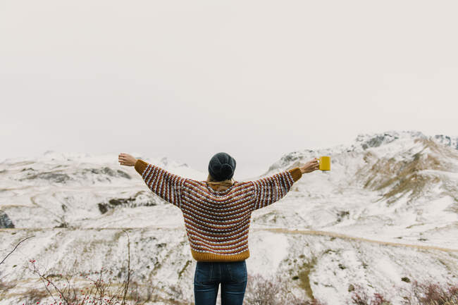 Повернення до молодої леді у светрі та капелюсі, що тримає кухоль з руками до боків біля пагорбів у снігу в Піренеях. — стокове фото