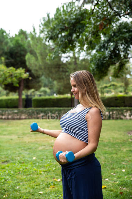Вид сбоку беременная женщина в спортивной одежде делает упражнения с гантелями в зеленом парке — стоковое фото