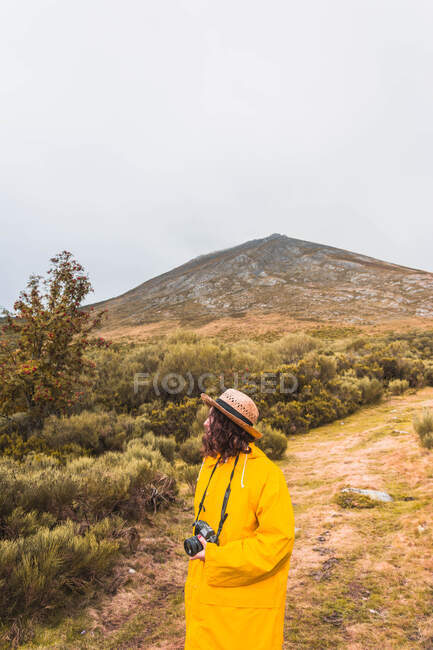 Vista lateral da senhora de chapéu e capa de chuva amarela com câmera olhando para a paisagem em Isoba, Castela e Leão, Espanha — Fotografia de Stock