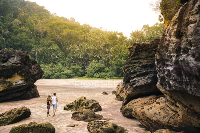 Vista posterior joven pareja entre rocas en la playa de arena cerca de bosque tropical verde en Malasia - foto de stock