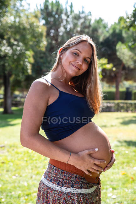 Вид сбоку молодая веселая беременная женщина в юбке и майке держа живот в зеленом саду — стоковое фото