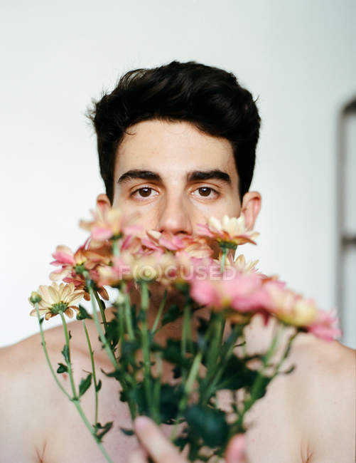 Morena joven sin camisa chico sosteniendo rosa flores frescas mirando a la cámara sobre fondo gris - foto de stock