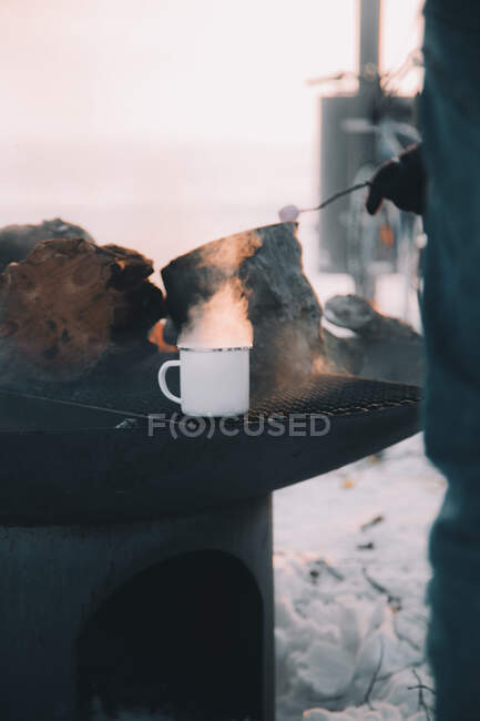Неузнаваемый человек, стоящий рядом с горячей печью с подогревом металлической чашки в заснеженной сельской местности Арктики — стоковое фото