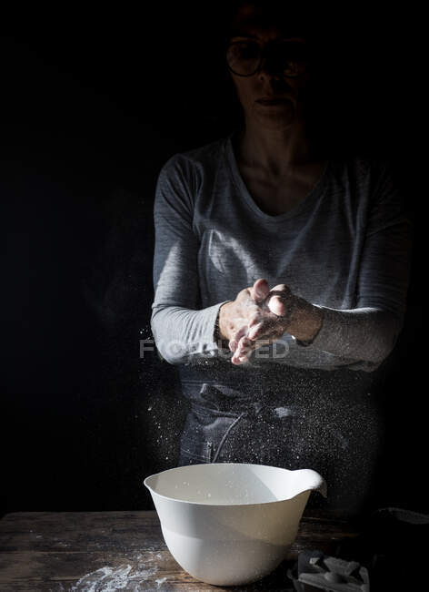 Женщина на полях хлопает в ладоши в муке возле миски, коробку с яйцами и взбивать на деревянный стол на черном фоне — стоковое фото
