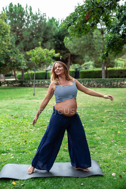 Веселая беременная женщина в спортивной одежде, растянувшаяся на коврике для упражнений в зеленом саду — стоковое фото