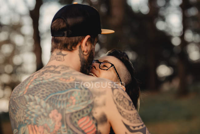 Hombre barbudo abrazando mujer alegre cerca de la madera en el bosque sobre fondo borroso - foto de stock