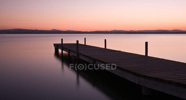 Longo cais de madeira perto do lago calmo durante o belo pôr do sol no campo incrível — Fotografia de Stock