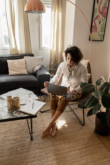 Frau mit Laptop auf Stuhl in der Nähe niedrigen Tisch mit Bleistiften, Pinseln und Papier im Zimmer — Stockfoto