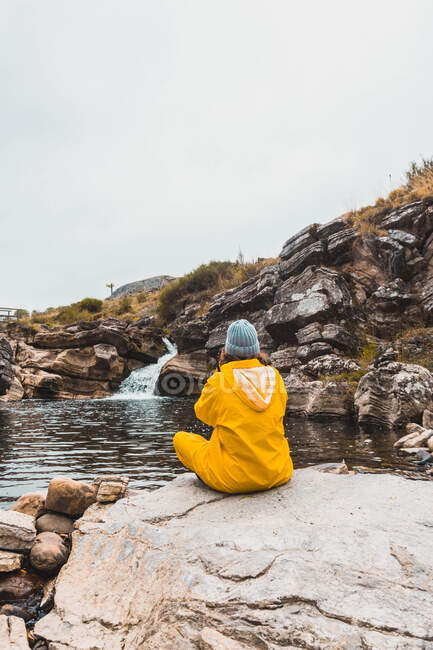 Back view of woman in hat and yellow raincoat sitting in stones and shooting rift of river between rock hills on camera in Isoba, Castile and Leon, Spain — Stock Photo