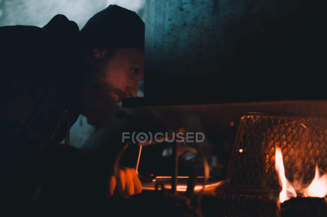 Seitenansicht eines bärtigen Typen, der in einen brennenden Ofen in einem kalten arktischen Gebäude blickt — Stockfoto