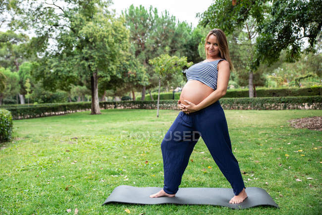 Усміхнена вагітна приваблива жінка тренується на килимку в парку — стокове фото