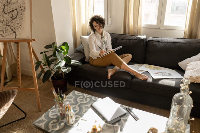 Жінка пише на папері на дивані в кімнаті. — стокове фото