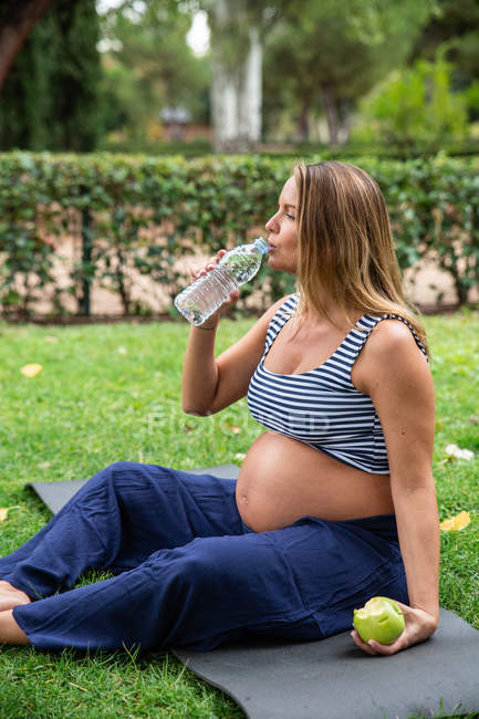 Pregnant attractive woman with water and apple on mat in park — Stock Photo