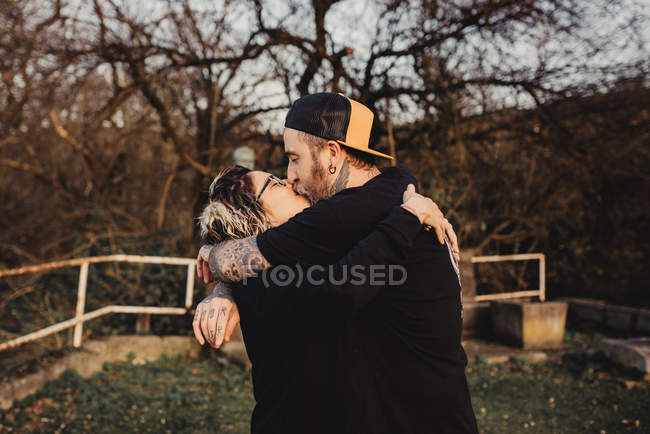 Боковой вид бородатого мужчины обнимающего и целующего женщину в очках в парке на размытом фоне — стоковое фото