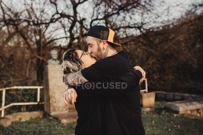 Боковой вид бородатого мужчины обнимающего и целующего женщину в очках в парке на размытом фоне — стоковое фото
