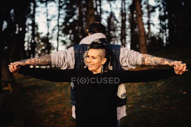 Jovem em tatuagens com pé de costas para trás e de mãos dadas com mulher sorridente no parque em fundo borrado — Fotografia de Stock