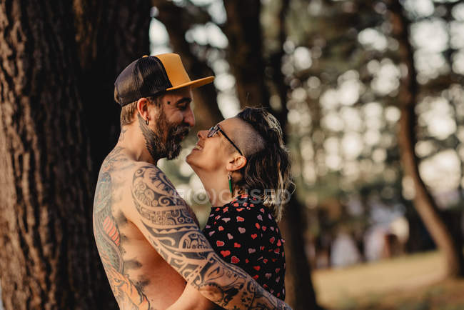 Giovane uomo senza maglietta in tatuaggi con abbraccio donna nel parco su sfondo sfocato — Foto stock