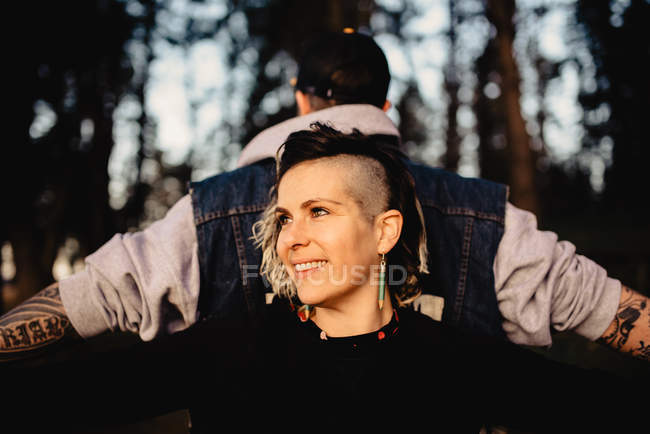 Young man in tattoos with standing back to back and holding hands with smiling woman in park on blurred background — Stock Photo