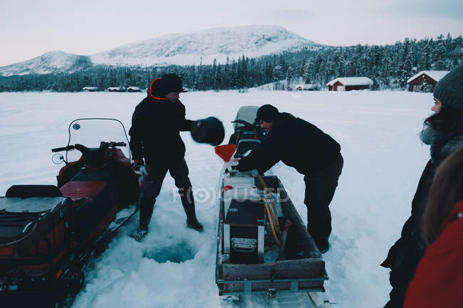 Deux jeunes hommes mettent de l'équipement sur des motoneiges modernes dans une magnifique campagne arctique par une froide journée d'hiver — Photo de stock