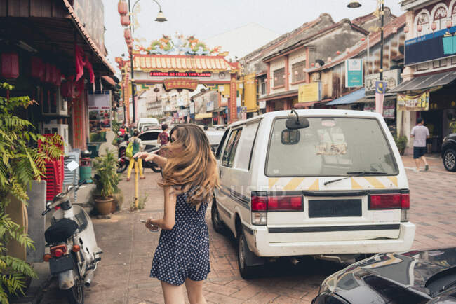 Back view young slim lady with windy hairs going on Asian street between buildings and automobiles in Malaysia — Stock Photo