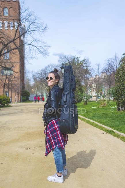 Vista lateral de una joven mujer hipster sonriente caminando en un parque en un día soleado mientras lleva una guitarra en la espalda - foto de stock
