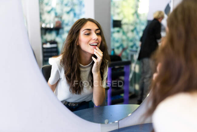 Reflejo de la joven hermosa dama con la mano cerca de los labios mirando el espejo en el salón de peluquería - foto de stock