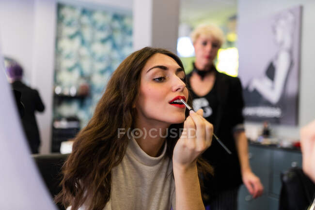 Riflessione di giovane bella signora con mano vicino alle labbra guardando lo specchio nel salone di parrucchiere — Foto stock