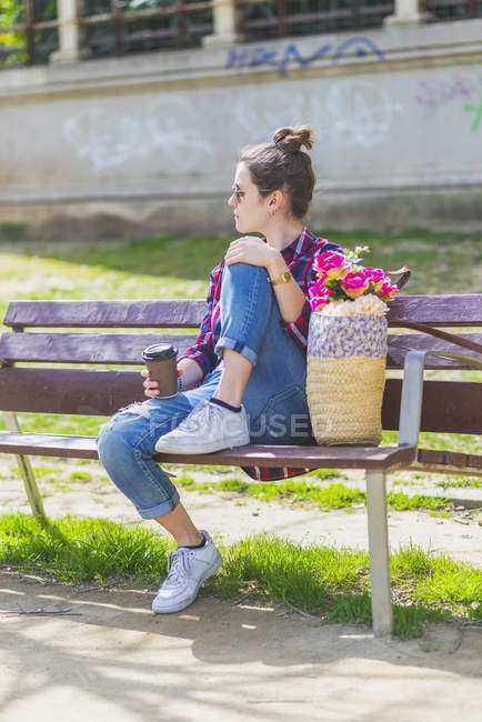 Vista lateral de una joven mujer hipster sentada en un banco del parque relajándose en un día soleado mientras mira hacia otro lado - foto de stock