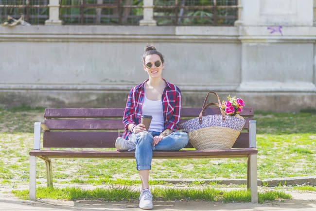 Vista frontal de una joven mujer hipster sentada en un banco del parque relajándose en un día soleado mientras mira la cámara - foto de stock
