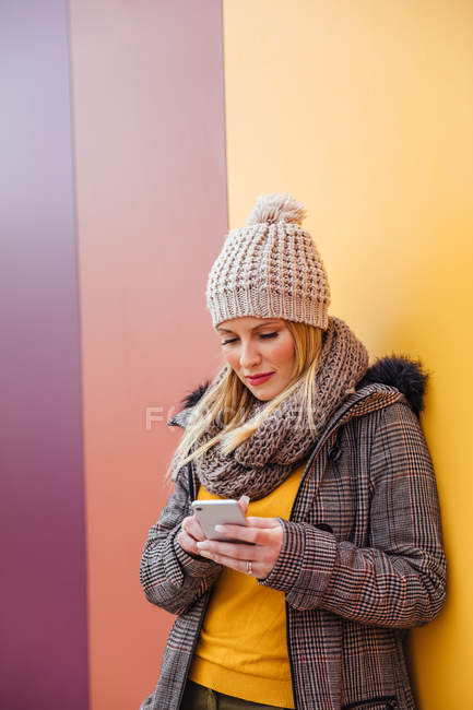 Portrait of blonde girl writing on her phone leaning against a colorful wall — Stock Photo