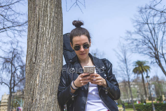 Вид сбоку на молодую смеющуюся хипстершу, стоящую и опирающуюся на дерево в парке в солнечный день, используя мобильный телефон — стоковое фото