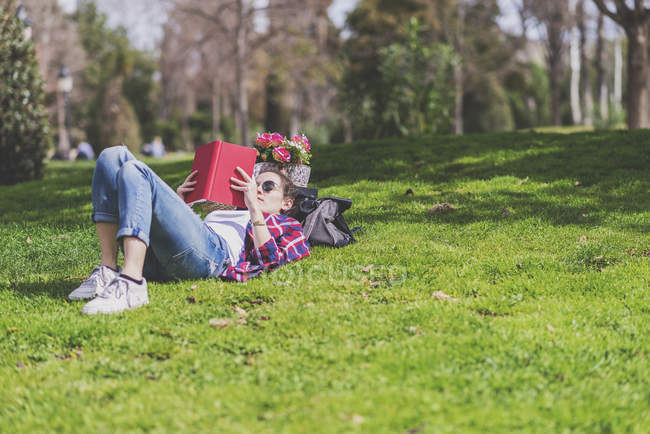 Vista lateral de una mujer feliz hipster acostado en la hierba en el día soleado en el parque mientras lee un libro rojo - foto de stock