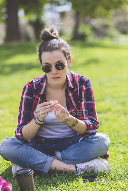 Vista frontal de uma jovem mulher hipster sentada na grama em um parque enquanto segura uma flor em um dia ensolarado — Fotografia de Stock