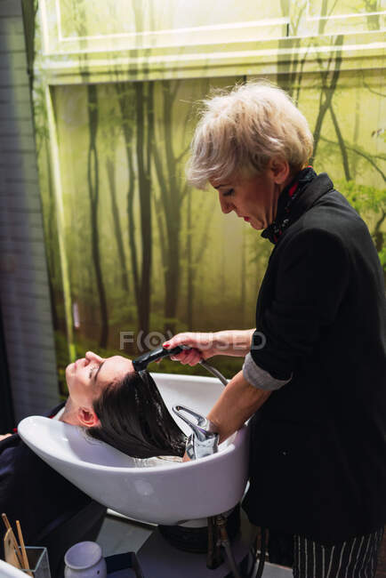 Aged stylist washing hairs of attractive lady near sink in hairdressing salon — Stock Photo