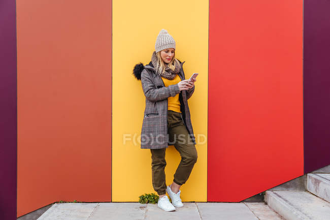 Portrait de fille blonde écrivant sur son téléphone appuyé contre un mur coloré — Photo de stock
