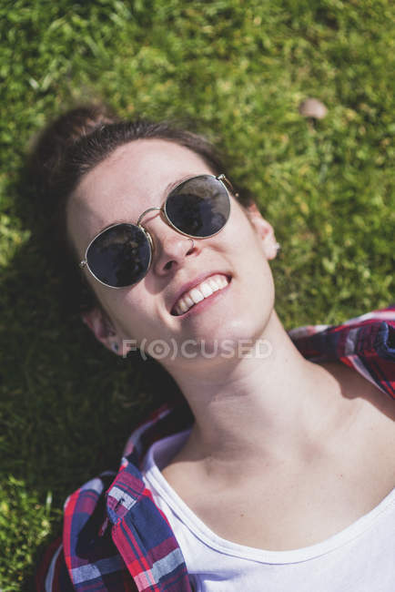 Над видом молодой улыбающейся хипстерши, лежащей на траве в солнечный день в парке, делая селфи с мобильным телефоном — стоковое фото