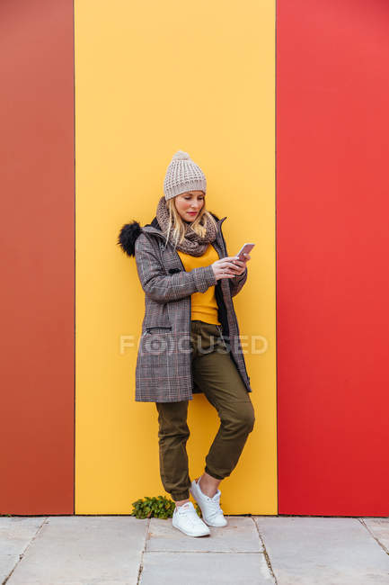 Porträt eines blonden Mädchens, das an eine bunte Wand gelehnt auf ihr Handy schreibt — Stockfoto