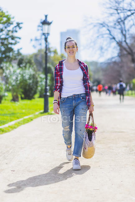 Вид на молодую улыбающуюся хипстершу, гуляющую в парке в солнечный день, держа в руках злую корзину — стоковое фото