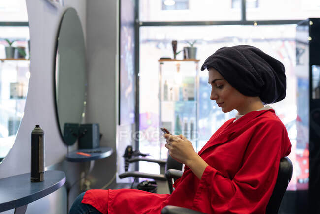 Vista lateral de la atractiva dama con toalla en la cabeza sentado en la silla en el salón de peluquería y la navegación en el teléfono móvil - foto de stock