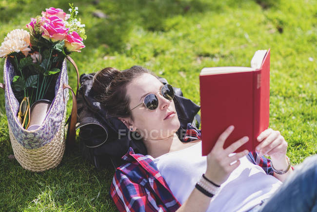 Vista lateral de uma mulher feliz hipster deitado na grama no dia ensolarado no parque enquanto lê um livro vermelho — Fotografia de Stock