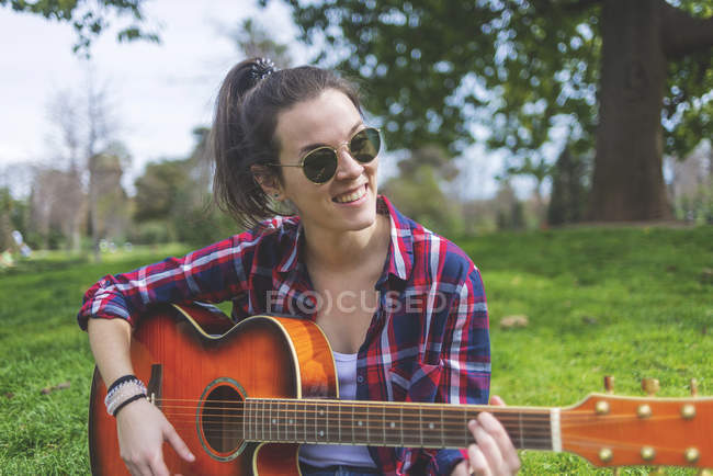 Вид спереди на молодую хипстершу в солнечных очках, сидящую на траве в парке и играющую на гитаре — стоковое фото